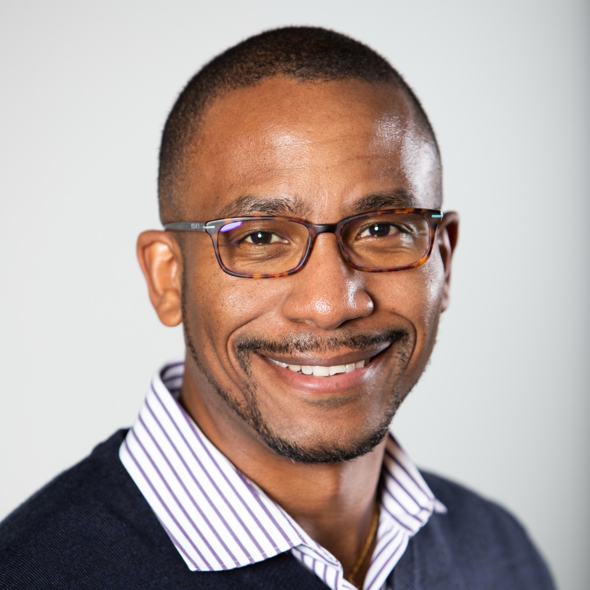 Johnny Boursiquot smiling, in glasses, a striped collar, and a navy sweater